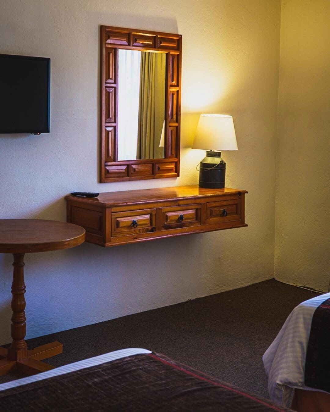 A hotel room with a wooden dresser, a table, a lamp, and a TV mounted on the wall.