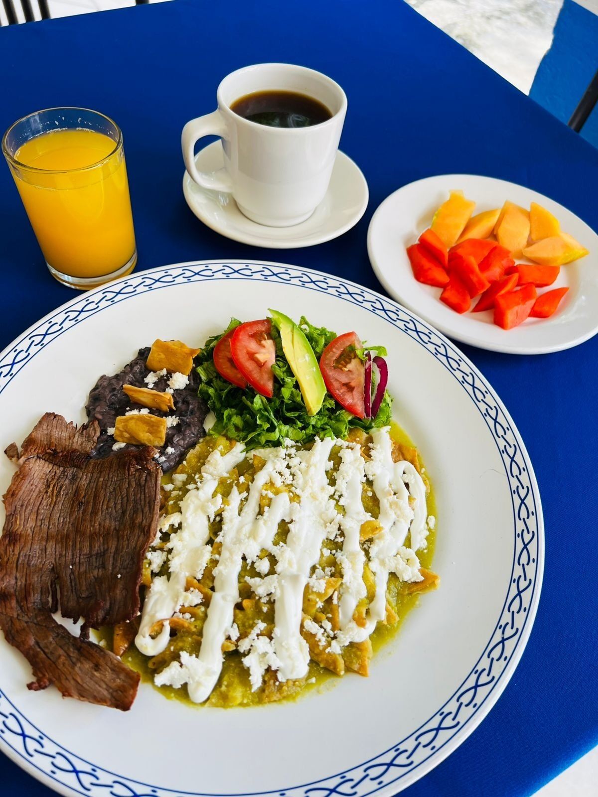 A plate of food with a side of fruit, a glass of orange juice, and a cup of coffee on a blue table.