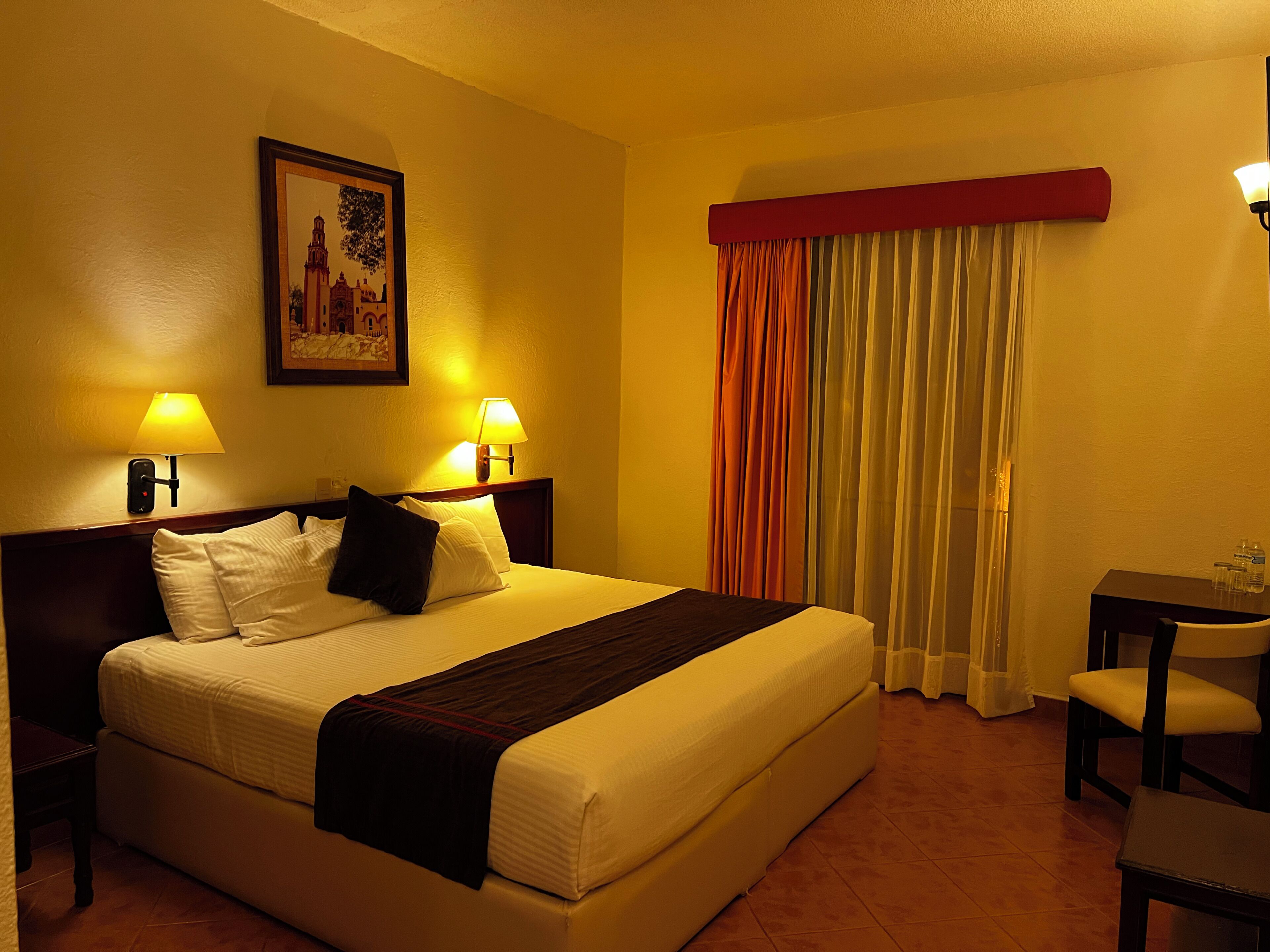 Habitación de hotel con cama, dos lámparas de noche, un cuadro en la pared, una ventana con cortinas y una mesita con una silla.