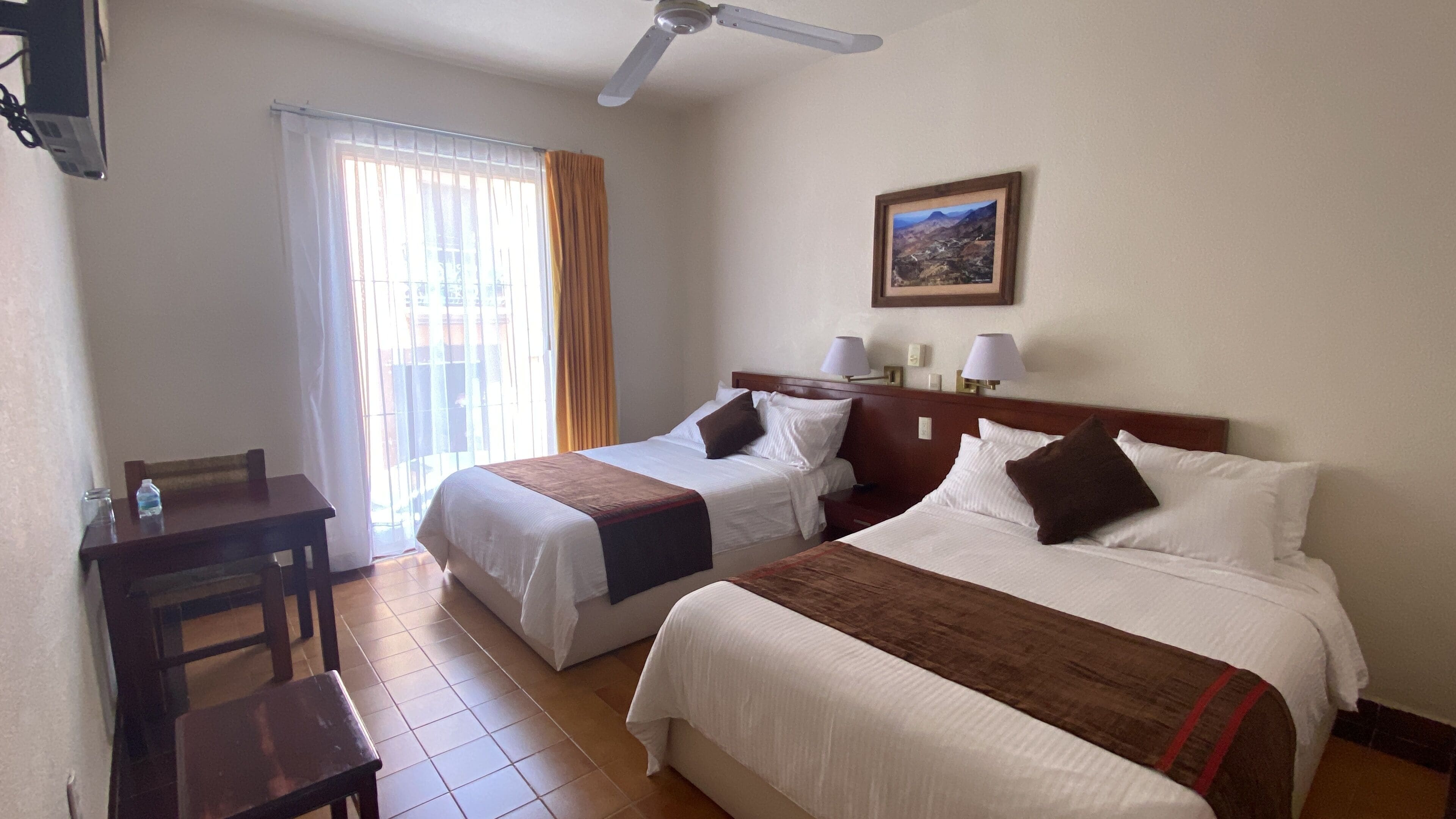 Una habitación de hotel con dos camas, un escritorio, una silla, un ventilador de techo y una ventana con cortinas.