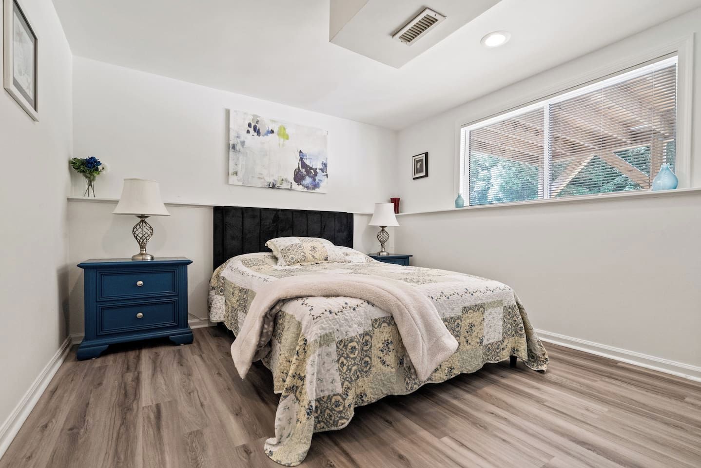 Une chambre à coucher comprenant un lit, deux tables de chevet, une œuvre d’art accrochée au mur, une fenêtre avec des stores et une bouche d’aération au plafond.