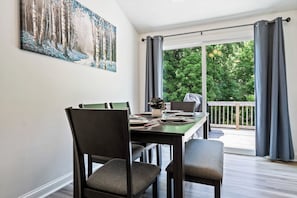 A dining area with a table set for four, chairs, a painting on the wall, and a view of greenery through large windows.