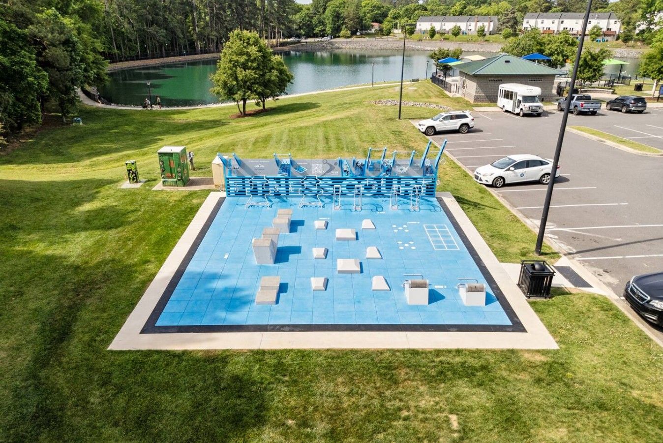 A swimming pool with a blue deck, surrounded by grass and a parking area with vehicles.