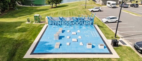 Una piscina con suelo azul, rodeada de césped y un aparcamiento con vehículos.