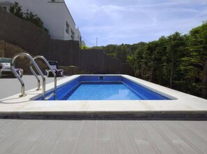 Une piscine rectangulaire avec un escalier en métal, entourée d’une terrasse en bois et d’un bâtiment blanc.