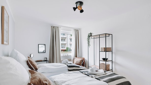 Una habitación de hotel moderna con dos camas, una mesita, una silla, una planta colgante y vistas a la ciudad.