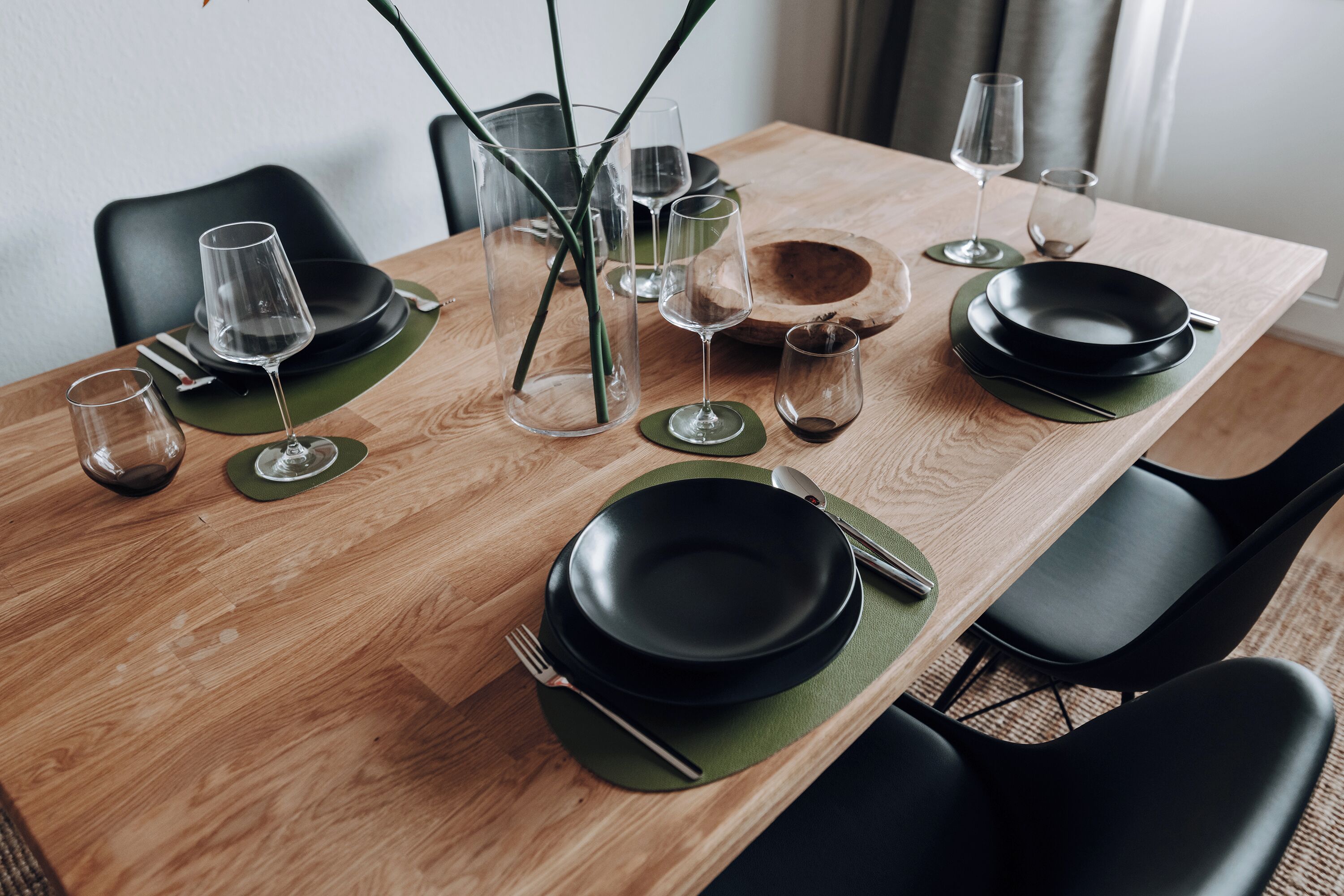 A wooden dining table set with black plates, clear glasses, and green placemats.
