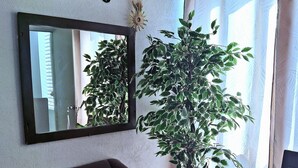 Ein Wohnzimmer mit einer Couch, einem grün-weißen Teppich, einer großen Kübelpflanze, einem Spiegel und einem Fenster mit Vorhängen.
