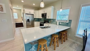 A modern kitchen with white cabinets, a large island with stools, stainless steel appliances, and a view of the outdoors through a window with blinds.