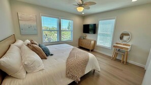 Une chambre à coucher avec un lit, un ventilateur de plafond, une télévision, une commode et deux fenêtres.