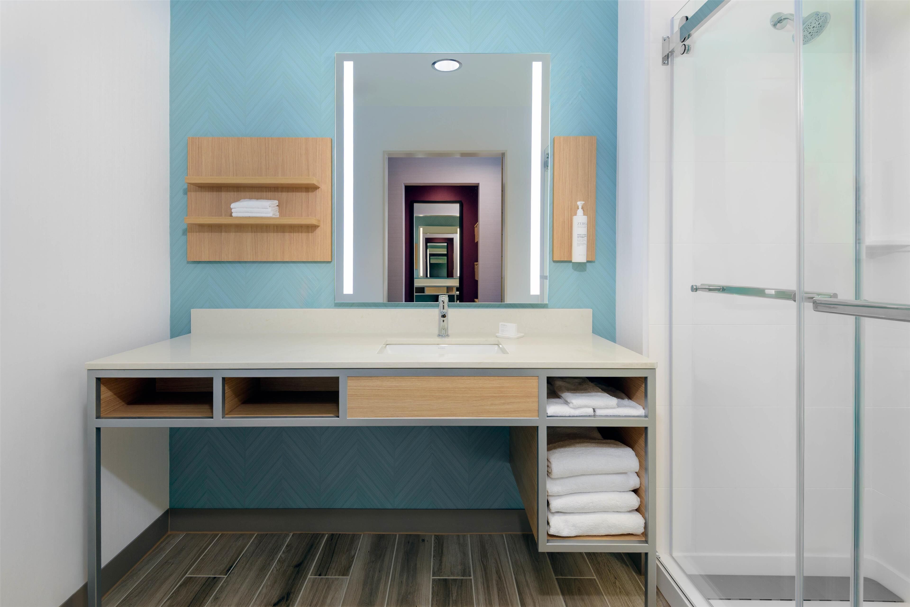 Une salle de bain moderne avec un grand miroir, un lavabo et une douche. On trouve des étagères en bois au-dessus du lavabo et un porte-serviettes sur la droite.