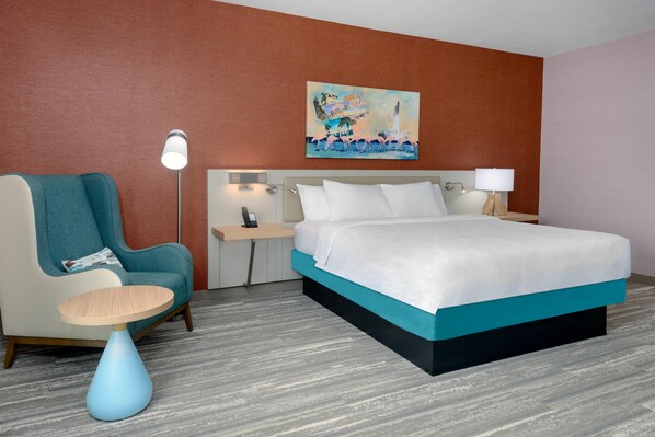 Habitación de hotel con cama, una silla, mesita de noche, lámpara y un cuadro en la pared.