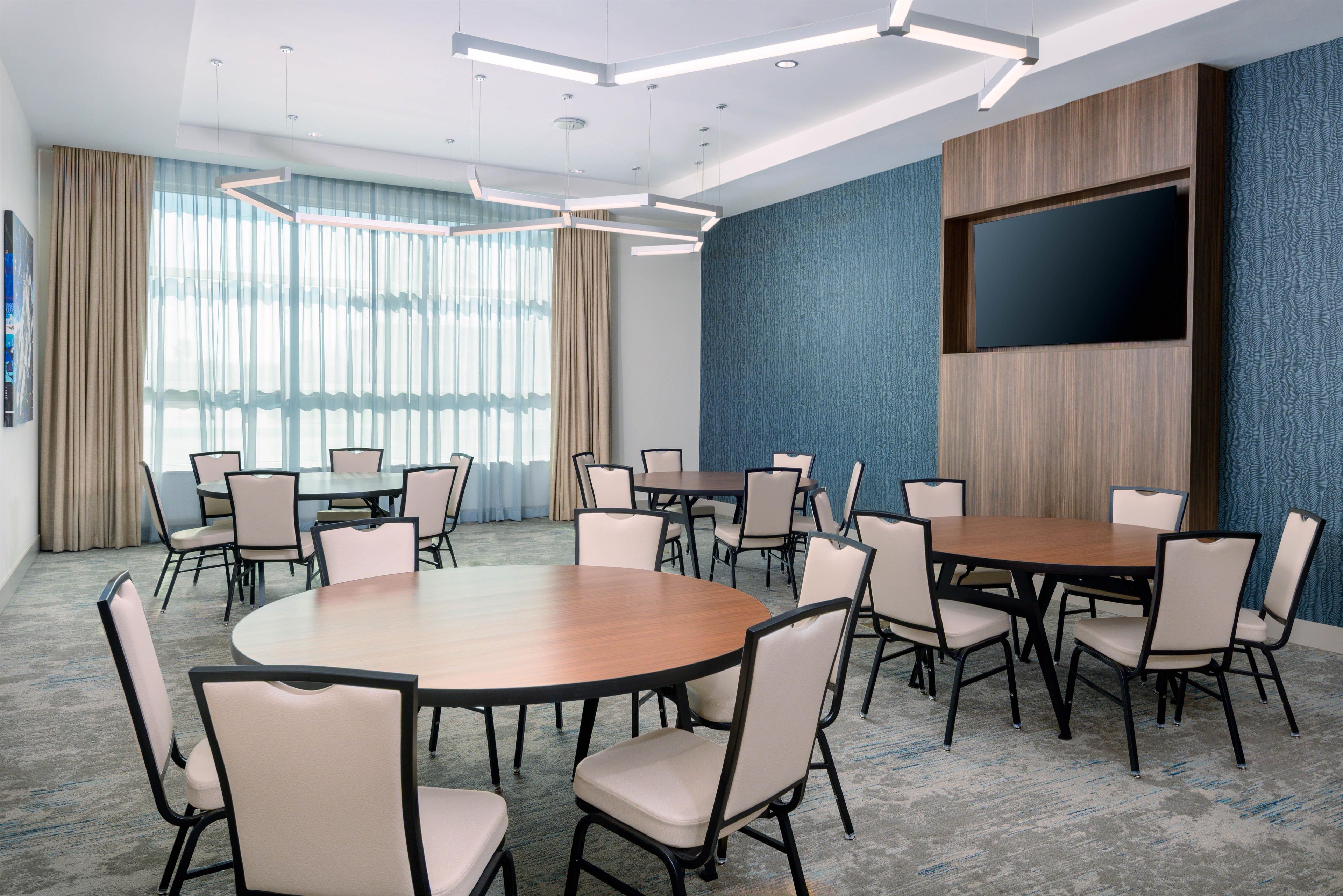 A conference room with a large table and chairs, a flat-screen TV mounted on the wall, and large windows with curtains.