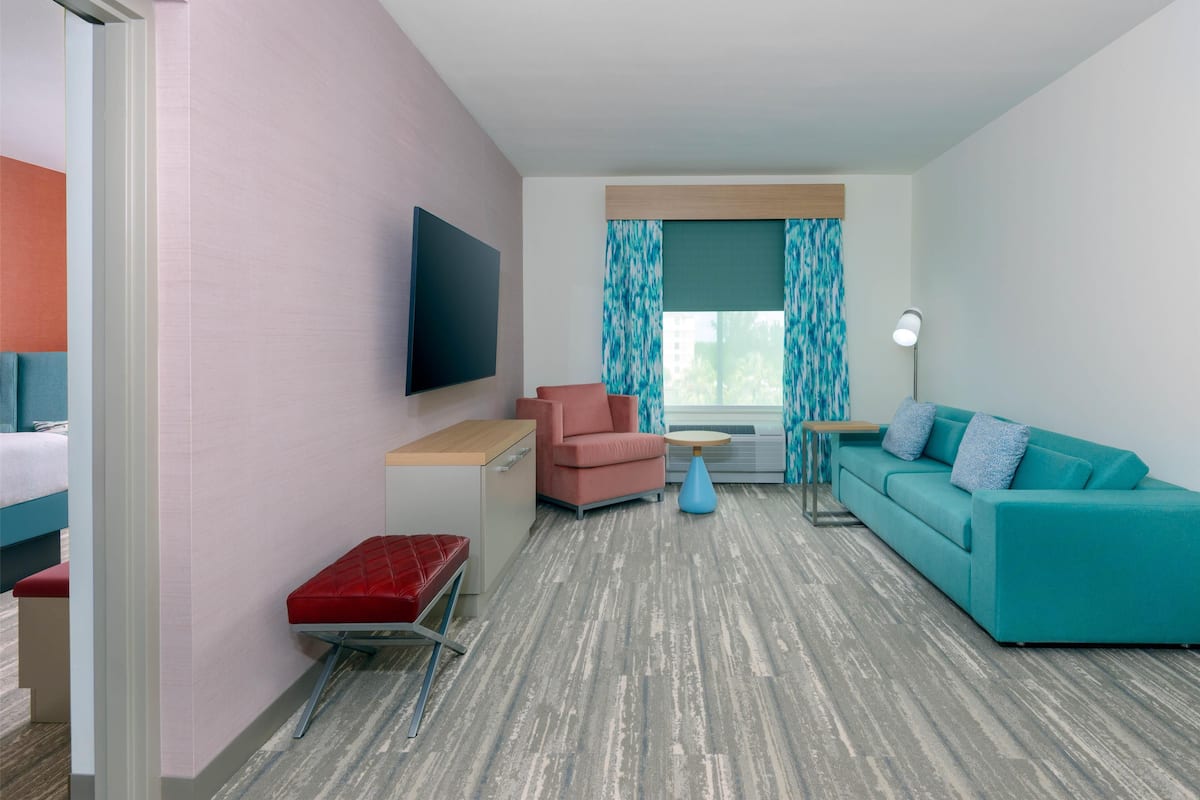 Une chambre d’hôtel moderne avec un canapé turquoise, un banc rouge et un téléviseur à écran plat fixé au mur.