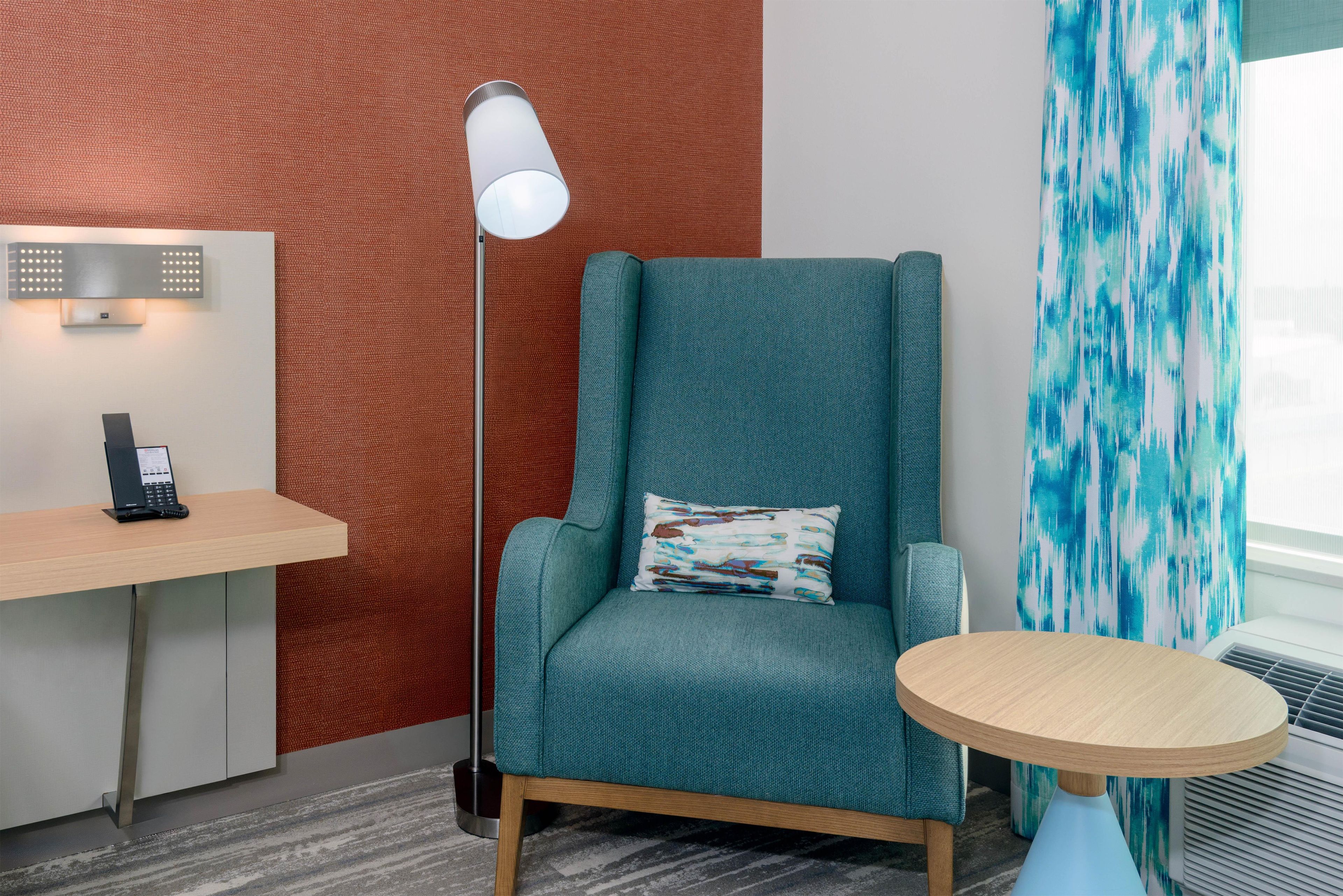 A modern room with a teal armchair, a small round table, a desk with a phone, a floor lamp, and a patterned curtain.