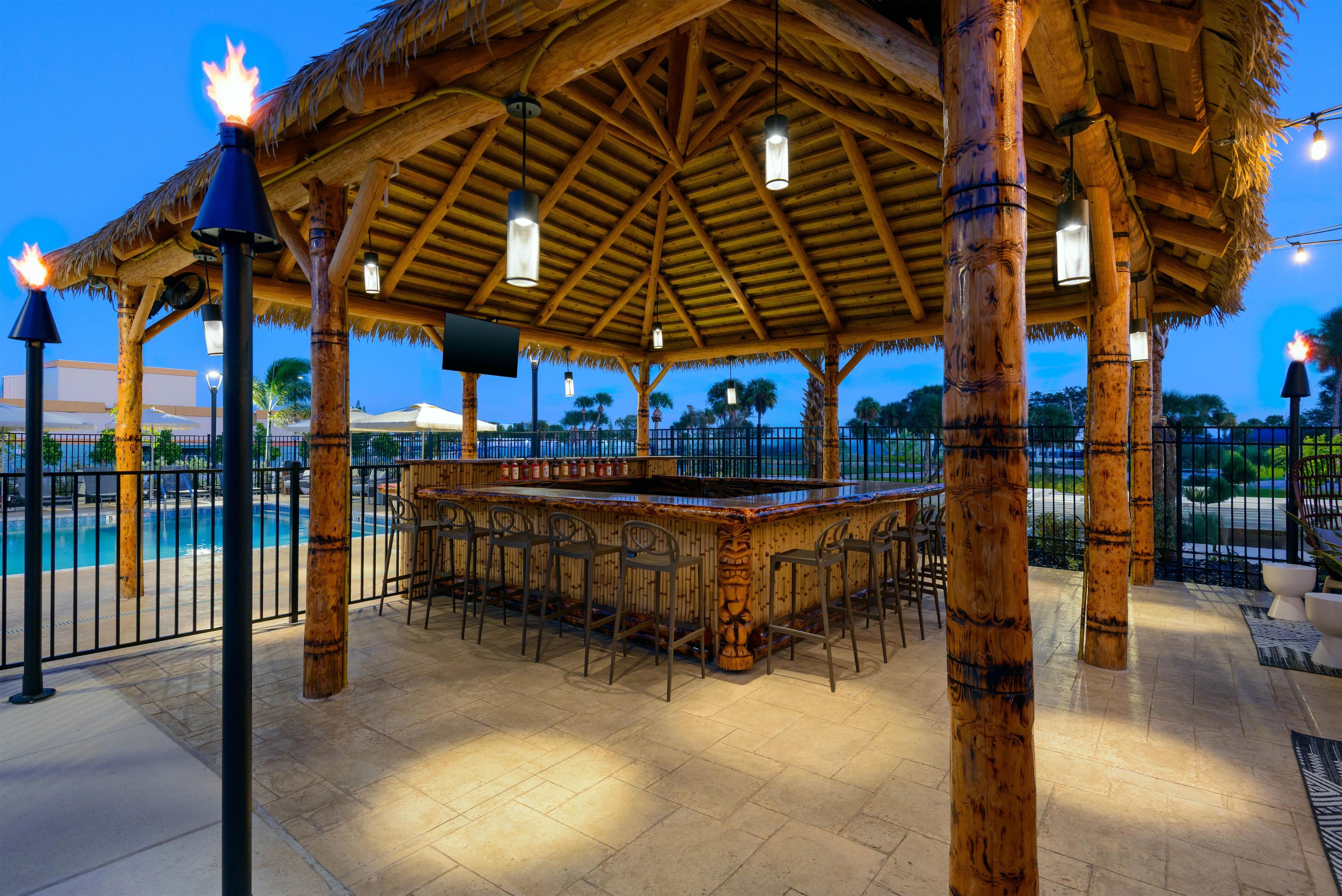 A tiki bar with a thatched roof, surrounded by a pool and palm trees.