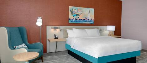 A hotel room with a bed, a chair, a desk, a lamp, and a painting on the wall.