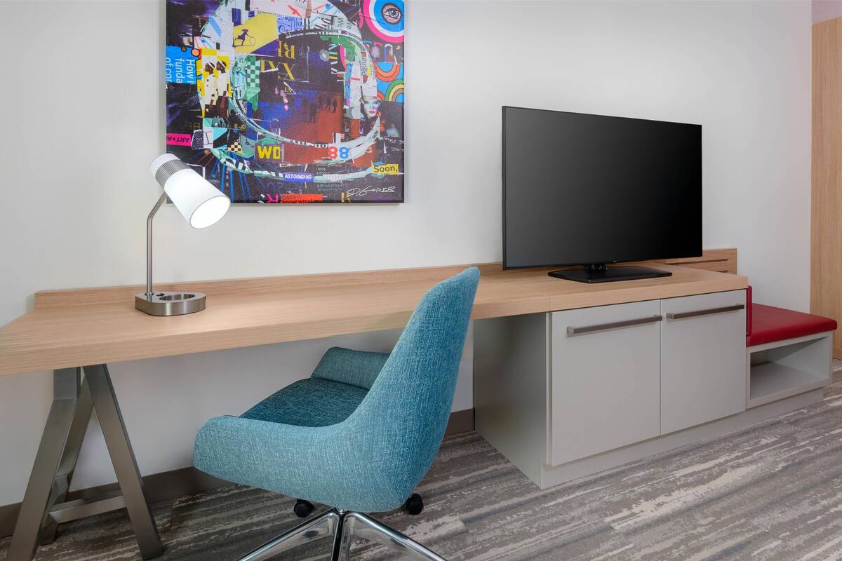 Un espace de bureau moderne comprenant un bureau, une chaise bleue, un téléviseur et un tableau abstrait aux couleurs vives accroché au mur.