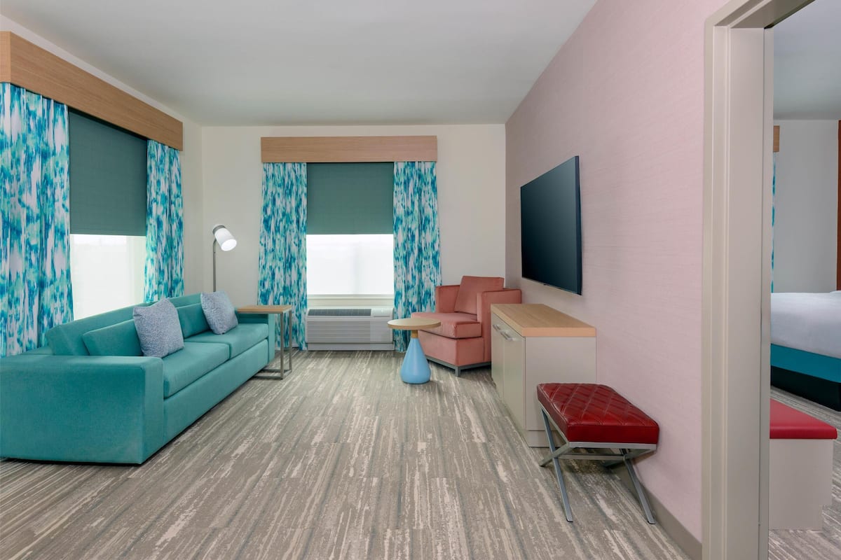Une chambre d’hôtel avec un canapé de couleur turquoise, un banc rouge, une petite table, un téléviseur à écran plat et un lit.