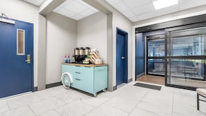 A clean, modern lobby with a blue door, a blue cart with amenities, and a large glass door leading outside.