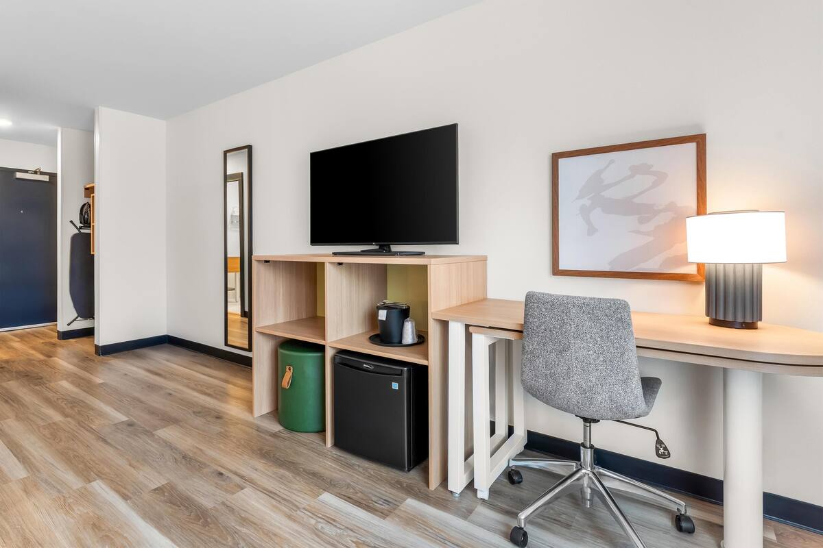 Quarto de hotel moderno com televisão de tela plana, escrivaninha com luminária, cadeira, mini-geladeira e lixeira.