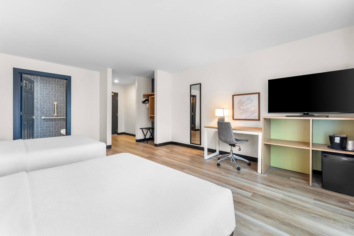 Quarto de hotel com cama, escrivaninha, cadeira, TV e cozinha compacta.