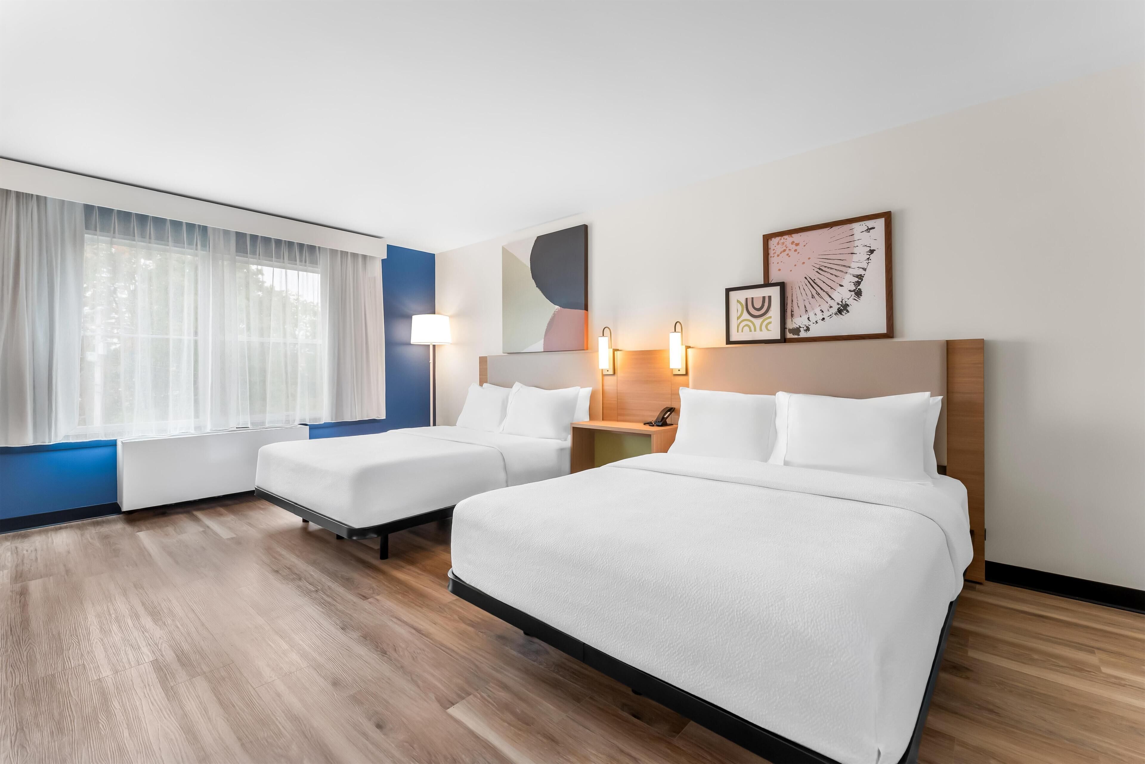 Een hotelkamer met twee bedden, een groot raam met dunne gordijnen, een blauwe accentmuur en kunst aan de muur.
