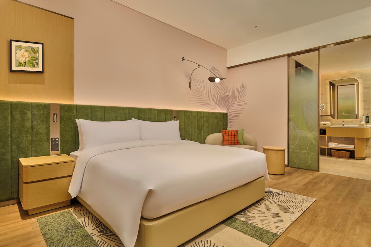 A modern hotel room with a large bed, a nightstand, a bedside table, a wall-mounted lamp, and a bathroom visible through a sliding door.