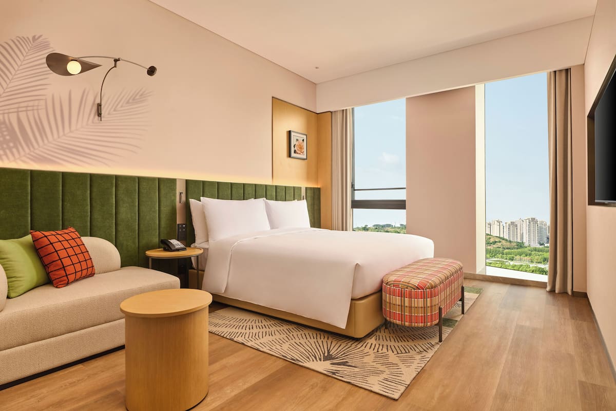 A modern hotel room with a large bed, a sofa, a round side table, and a view of a cityscape through large windows.