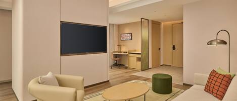 A modern living room with a sofa, armchair, and coffee table. There is a desk with a chair and a television mounted on the wall.