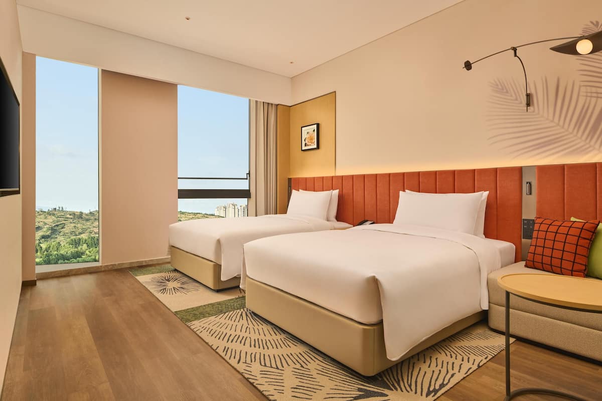 A modern hotel room with two beds, a large window with a city view, a small table, and a mounted TV.