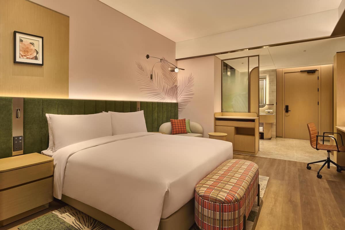 A modern hotel room with a large bed, a desk, a chair, a mirror, and a small bathroom.