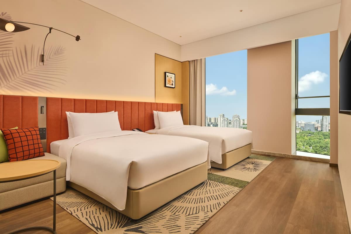 A modern hotel room with two beds, a small table, a sofa, a large window with a city view, and a ceiling fan.