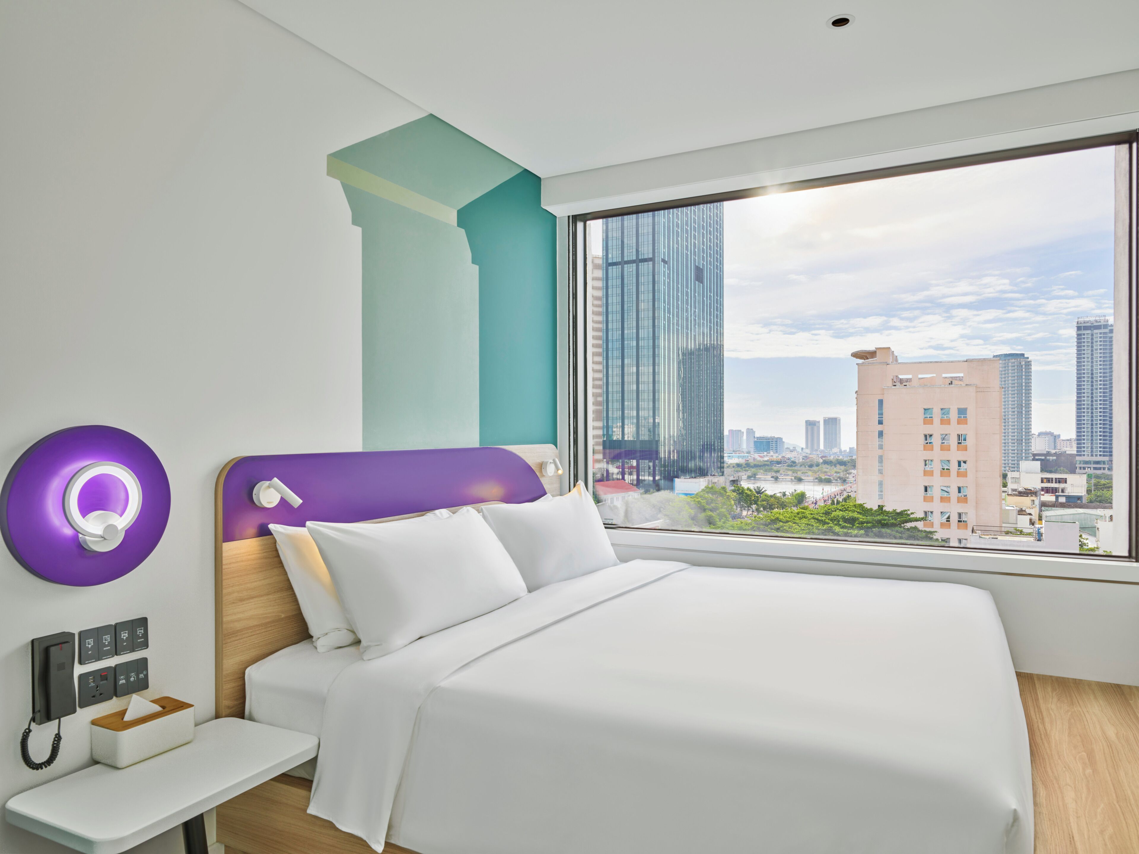 Une chambre d’hôtel moderne avec un grand lit, une table de chevet dotée d’un téléphone et de mouchoirs, un mur d’accent violet et une vue sur la ville par une grande fenêtre.