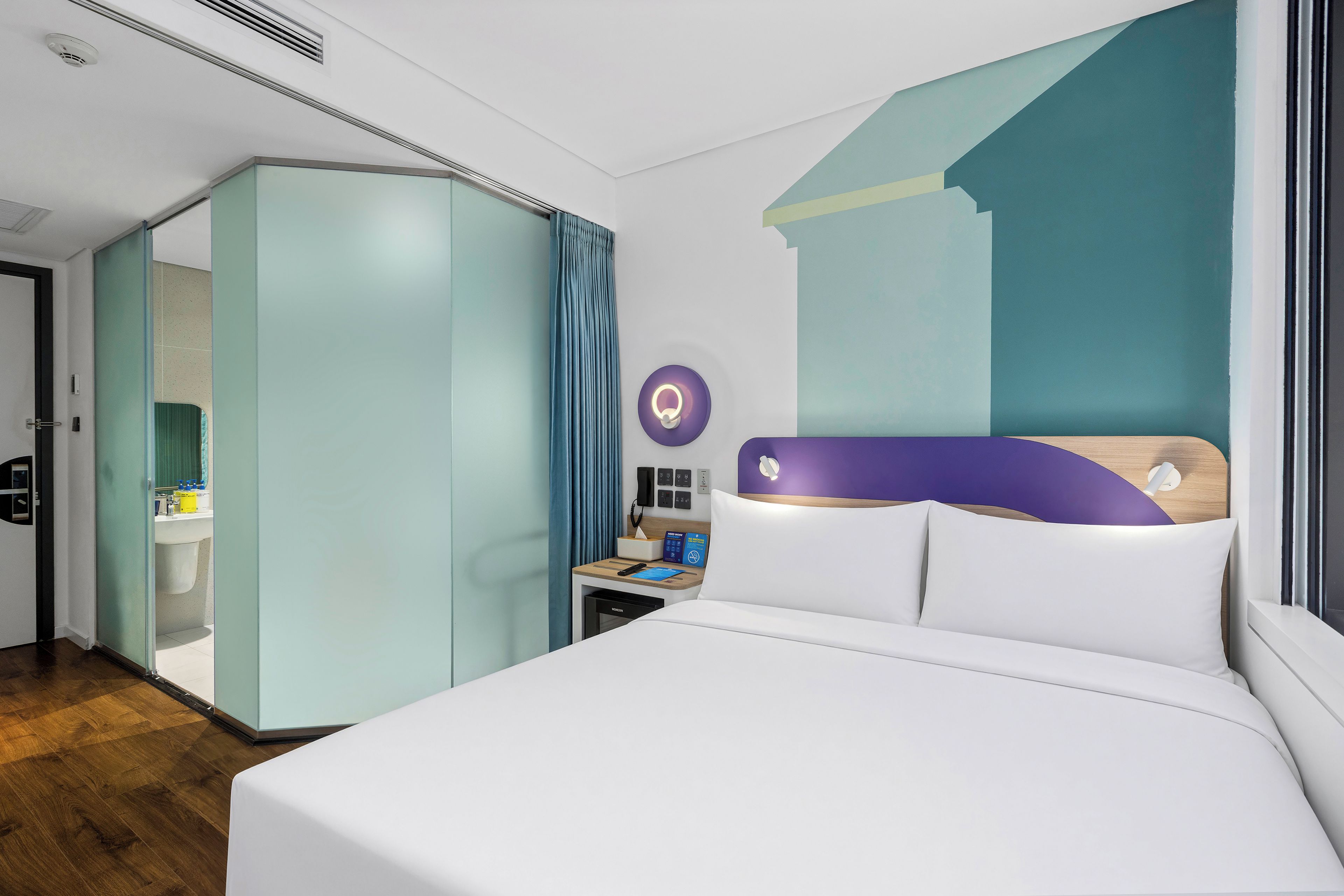 Ein modernes Hotelzimmer mit einem großen Bett, einem Kleiderschrank, einem Nachttisch mit Lampe und einem durch eine Glaswand abgetrennten Badezimmer.