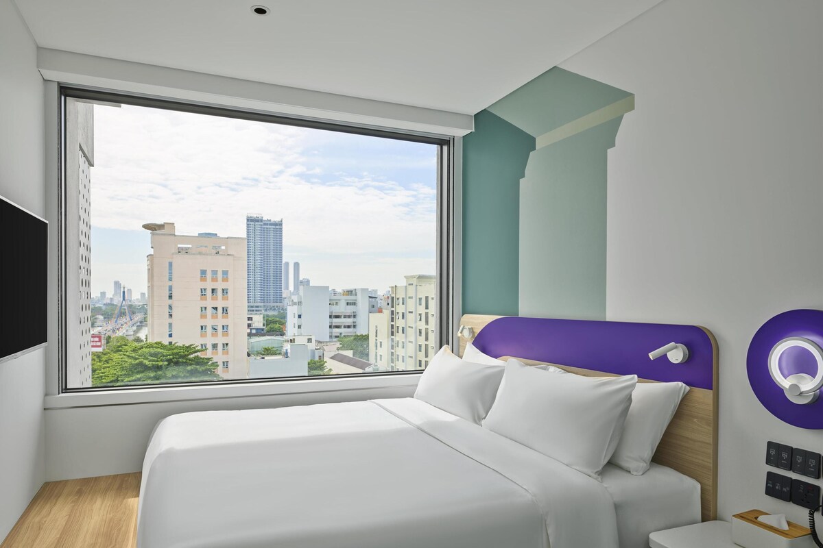 Una habitación de hotel moderna con un ventanal grande, una cama con ropa blanca, piso de madera y una pared de acento color púrpura.