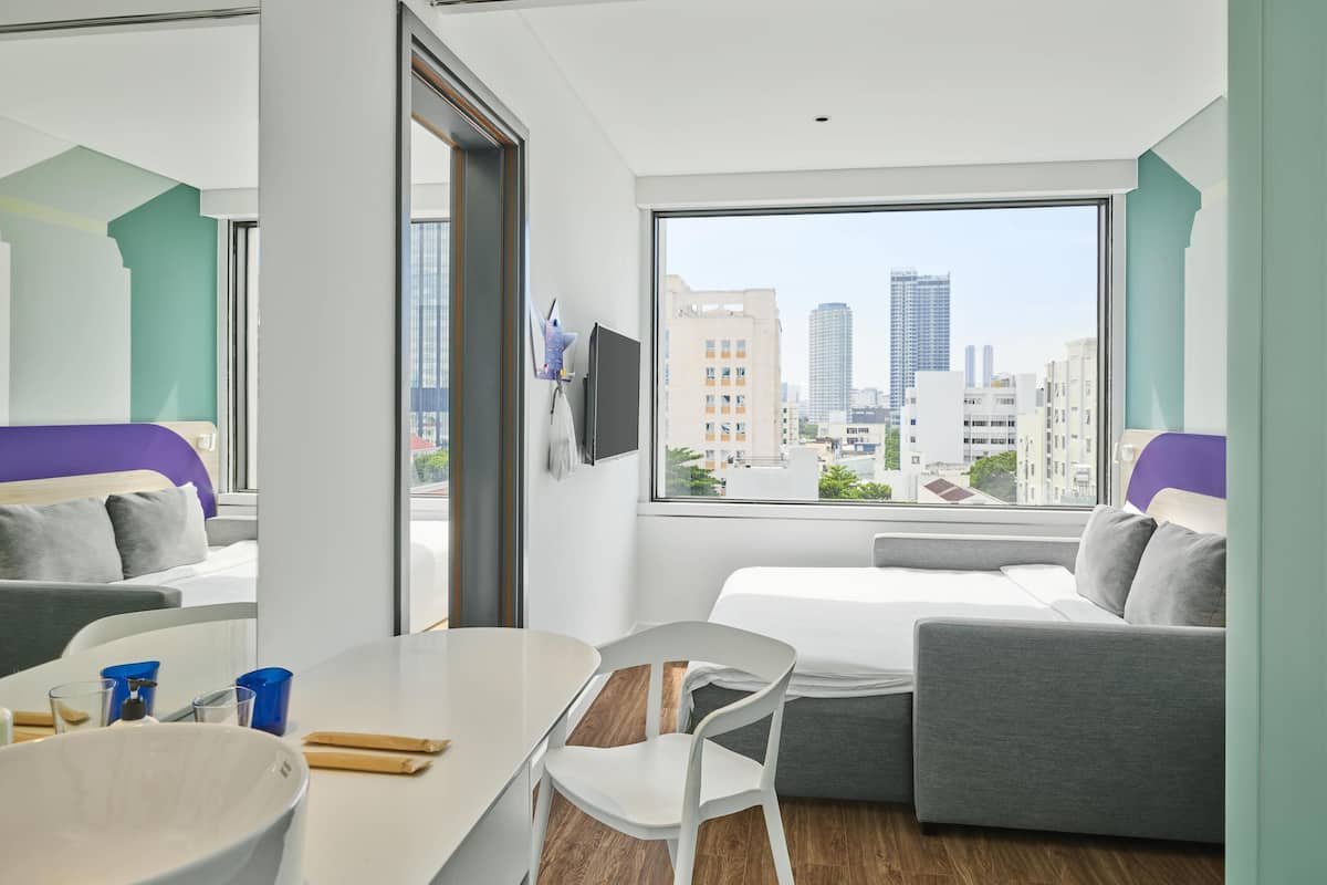 A modern hotel room with a large window, a bed, a sofa, a dining table, and a TV.