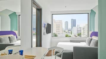 A modern hotel room with a large window, a bed, a sofa, a dining table, and a TV.