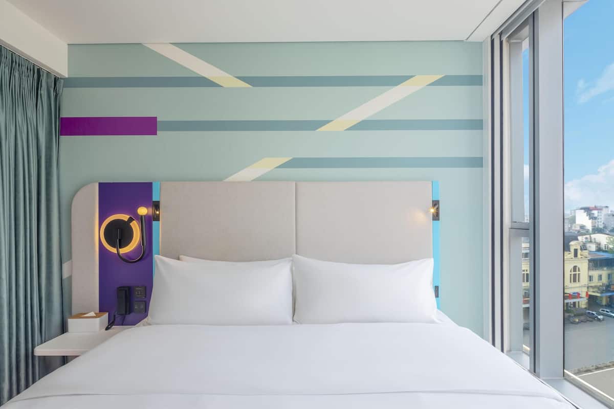A modern hotel room with a large bed, a nightstand, a wall-mounted phone, and a window with a city view.
