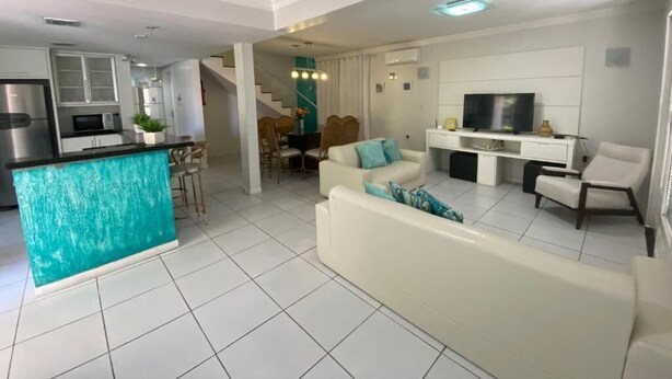 A modern living room with a white sofa, a kitchen area with a turquoise island, and a dining area with chairs and a table. - Duplex with barbecue 70 meters from the sea on Ingleses beach (Ingleses Sul Florianópolis)