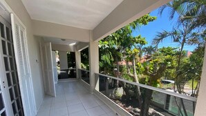 A balcony with a tiled floor, a railing, and a view of a garden with trees and a car. - Duplex with barbecue 70 meters from the sea on Ingleses beach (Ingleses Sul Florianópolis)