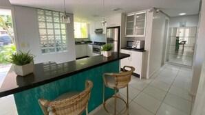 A modern kitchen with a teal island, white cabinets, and a black countertop. There are two wicker chairs and a potted plant on the island. - Duplex with barbecue 70 meters from the sea on Ingleses beach (Ingleses Sul Florianópolis)