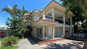 A two-story building with a covered patio area, a gravel driveway, and surrounding palm trees. - Duplex with barbecue 70 meters from the sea on Ingleses beach (Ingleses Sul Florianópolis)