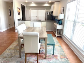 A dining area with a wooden table and white chairs, a kitchen with white cabinets, a refrigerator, and a window with blinds. - 4BR Fort Morgan: Stunning Gulf Views, Across From New Pool, 2 min Walk To Beach (Gulf Shores)