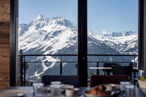 Espaço de refeições com vista para a montanha, com mesa, cadeiras e picos nevados.
