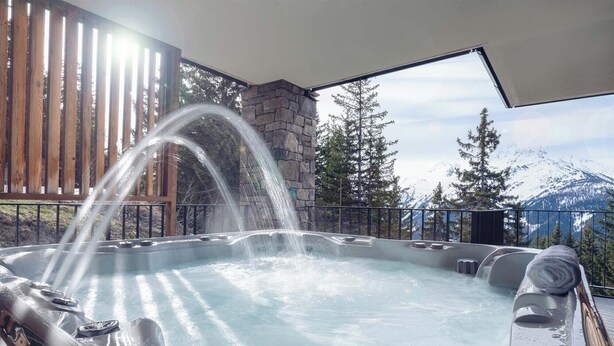 A hot tub with a waterfall feature, surrounded by a wooden deck and stone walls, with a mountain view in the background.