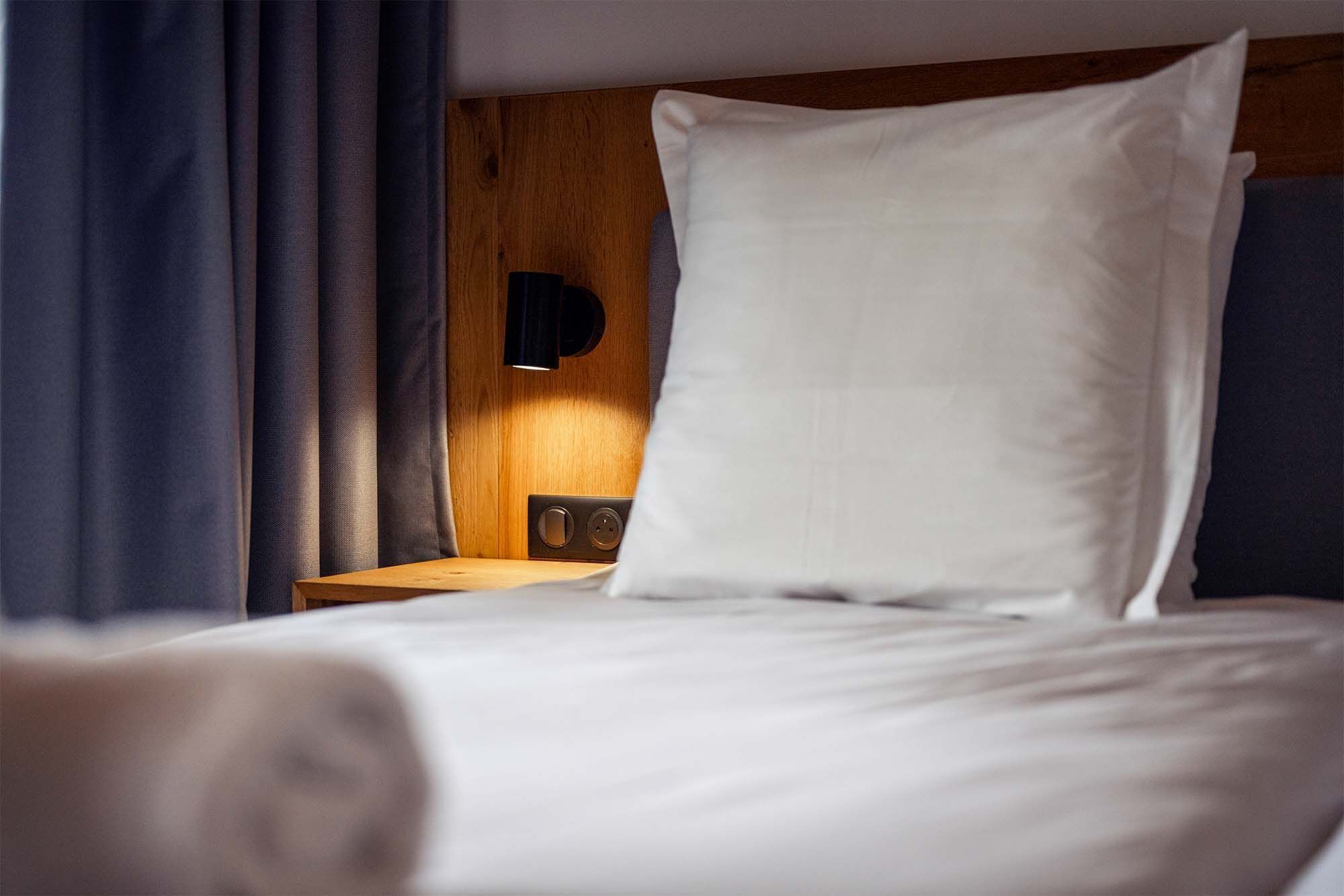 Un lit bien fait, avec du linge de lit blanc, une tête de lit en bois et une table de chevet surmontée d’une lampe.