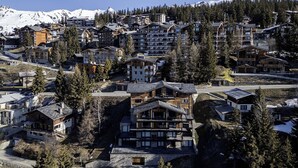 A mountainous resort area with multiple wooden buildings, some with balconies, surrounded by trees and snow-capped mountains in the background.