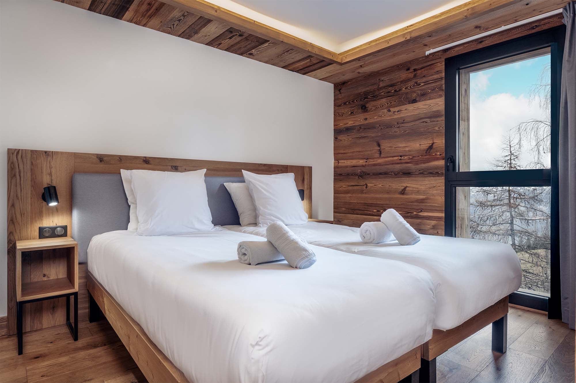 A modern bedroom with a wooden ceiling, a large bed with white linens, a bedside table with a lamp, and a window with a view of trees.