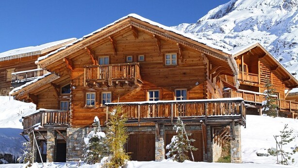 Un chalet en bois doté de plusieurs balcons et fondé sur des pierres, entouré de neige et de montagnes.
